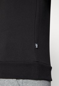 Primer plano del dobladillo de una sudadera negra con puños acanalados y una pequeña etiqueta del logo de Puma en el costado, combinada con pantalones grises.
