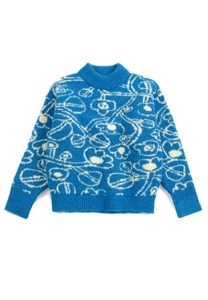 Jeune personne aux longs cheveux roux portant un pull bleu à motifs floraux, un pantalon jaune ample avec des détails colorés, et des bottes marron, debout les mains dans les poches.