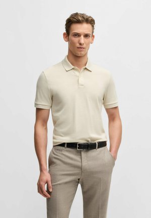 Polo beige en tissu doux, avec un col classique et des manches courtes, associé à un pantalon gris clair à motifs et une ceinture noire.