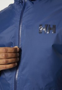 Blauwe waterdichte jas met een gestructureerde afwerking, voorzien van een zichtbare rits en een glanzend logo. Hand die de ritssluiting vastpakt voor aanpassing.