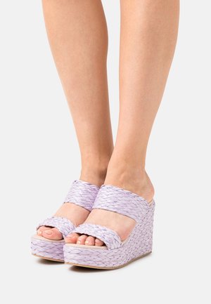 Gambe che indossano sandali con zeppa intrecciati di un leggero colore viola, con doppie cinghie, poggiate su una superficie bianca.