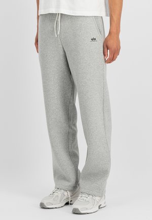 Uomo con pantaloni della tuta grigio chiaro con coulisse in vita e sneakers bianche, in piedi davanti a uno sfondo semplice.
