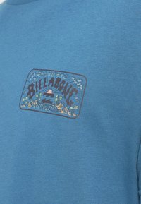 Blå bomulds-T-shirt med et logo-patch med farverige bølge- og palmetræsgrafikker på venstre brystområde. Kortærmet, rund hals.