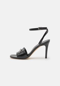 Black heeled sandal with a glossy finish, open toe design, ankle strap with buckle, and decorative rhinestone accent on the strap.