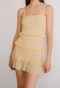 Žluté mini šaty s nabíraným designem, ozdobené volánkovými vrstvami a ramínky na tenké šňůrky; spodní vrstva má kostkovaný vzor, lehký materiál.