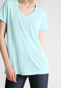 Kvinna iklädd en ljusblå V-ringad kortärmad skjorta och mörka leggings, stående med en hand avslappnad vid sin sida.