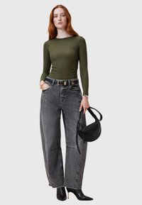 Maglietta verde oliva a maniche lunghe, jeans grigi a vita alta a gamba larga e tacchi a spillo neri. Borsa nera con un design curvo tenuta in mano.