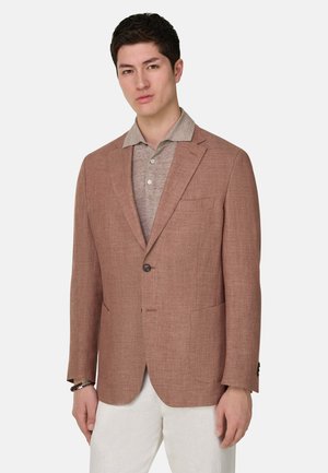 Mann trägt ein hellbraunes Sakko über einem beigen Poloshirt und weißen Hosen, steht vor einem einfarbigen Hintergrund.