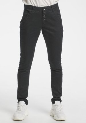 Pantalon noir slim avec quatre boutons visibles à l’avant porté avec des baskets blanches sur une personne debout.