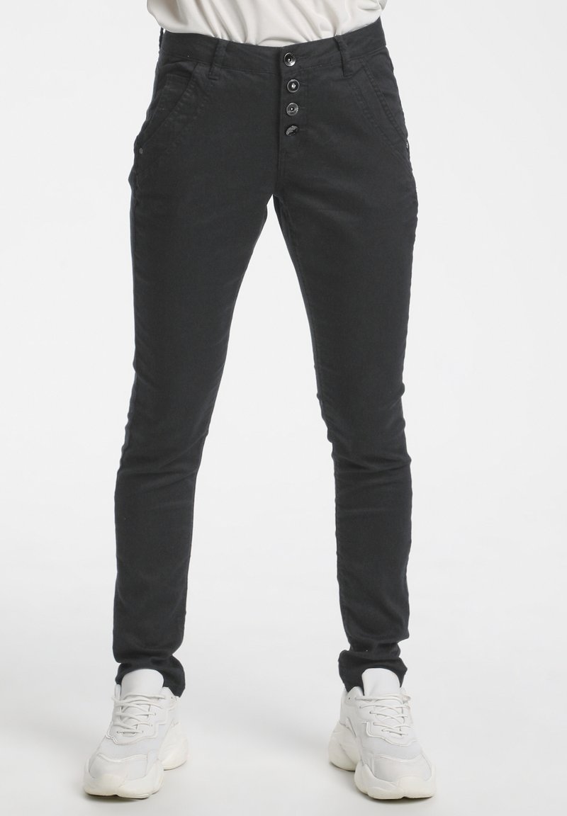 Pantalon noir slim avec quatre boutons visibles à l’avant porté avec des baskets blanches sur une personne debout.