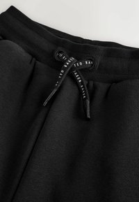 Mustad dressipüksid, millel on soonikvöökoht ja must nöör, millel on valges kirjas "Ted Baker".