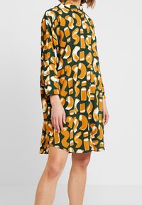 Långärmad klänning med krage i grön tyg som har orange och krämfärgade abstrakta former och mönster, med en avslappnad passform och längd till mitten av låren.