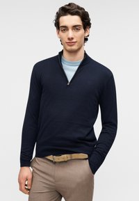 Pull-over à col zippé bleu marine en tissu fin tricoté, avec manches longues et texture lisse. Porté sur une chemise bleu clair avec un pantalon beige.