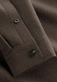 Braun strukturierter Hemdmanschetten mit einem schwarzen Knopf. Der Stoff hat eine weiche Oberfläche mit sichtbaren Nähdetails.