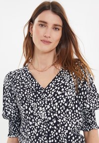 Sort bluse med hvide prikker, V-hals design, korte puffærmer og knaplukning. Glat stof med en lidt løs pasform.