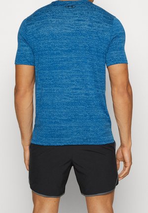 Man draagt een blauw sportief T-shirt met korte mouwen en zwarte hardloopshorts, gezien van achteren, staand tegen een effen achtergrond.