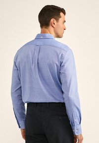 Camisa de manga larga azul claro hecha de tela texturizada, con un pliegue en la parte posterior, cuello tipo polo y acentos de puño abotonado.