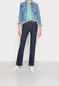 Hellblaues Denim-Jackett über einem grünen T-Shirt, kombiniert mit dunkelblauen Schlaghosen und weißen Sneakern, die ein lässiges Outfit präsentieren.