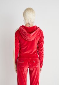 Punainen samettinen urheiluasu hupparilla, jossa on beigenväriset hihat. Kankaan pinta on sileä, ja takana on kaksi taskua metalliyksityiskohdilla.
