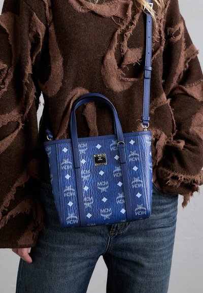 Tote bag en cuir bleu avec une surface à motifs présentant des logos MCM et des formes de diamant, des poignées doubles et une bandoulière réglable.