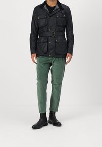 Tummansininen vyöllinen takki, jossa useita taskuja ja teksturoitu kaulus, yhdistetty vihreisiin samettihousuihin ja mustiin nilkkureihin.