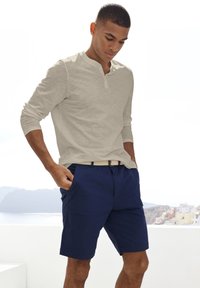 Maglietta a maniche lunghe color beige con colletto, abbinata a pantaloni corti blu navy. I pantaloni corti presentano tasche laterali e una cintura intrecciata color beige.