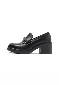 Mocassino in pelle nera con tacco spesso, caratterizzato da una prominente fascia decorativa e una finitura liscia, progettato per essere indossato facilmente.