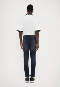 Λευκό κοντομάνικο πόλο μπλουζάκι με μαύρες λεπτομέρειες, φορεμένο με σκούρα μπλε στενά τζιν και μαύρα αθλητικά παπούτσια, αν viewed από πίσω.