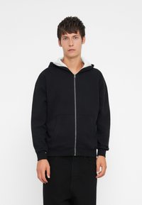 Fleece preto com capuz ajustável forrado a cinza, bolsos frontais e punhos canelados. O tecido parece texturizado e descontraído.