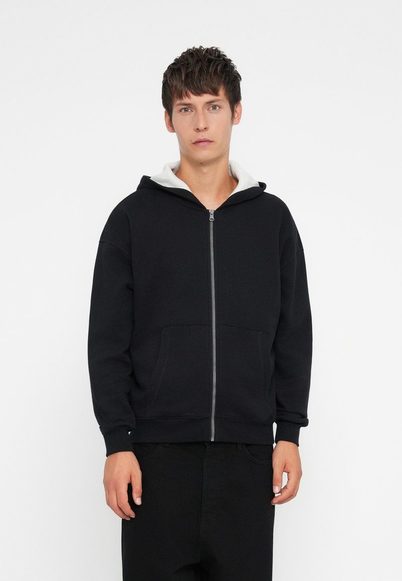 Fleece preto com capuz ajustável forrado a cinza, bolsos frontais e punhos canelados. O tecido parece texturizado e descontraído.