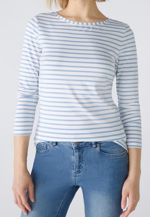 Femme portant un haut à manches longues à rayures horizontales blanches et bleu clair avec un jean en denim bleu, debout devant un fond uni.