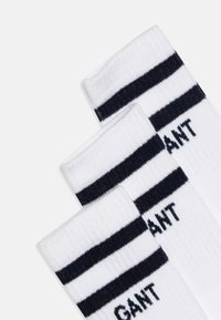 Vita ankelsockor med två svarta ränder nära toppen och "GANT" tryckt i svart på sidan. Mjukt material och ribbad Struktur.