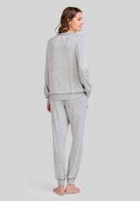 Mujer con cabello castaño vistiendo un top de manga larga gris claro con manchas y pantalones jogging a juego, de pie descalza, con las manos en los bolsillos, mirando hacia atrás.