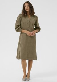 Olive green linen dress with a collar, button placket, and puffed sleeves. Hemline falls just above the knee; paired with light gray heels.
