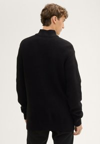 Schwarzer Strickpullover mit hohem Kragen, der ein strukturiertes Muster und einen lockeren Schnitt aufweist. Lange Ärmel reichen bis zu gerippten Bündchen.