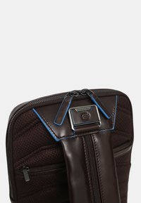 Bolso de cuero marrón con acentos azules, que presenta un diseño acolchado, cierres de doble cremallera y un cierre con logo para un ajuste seguro.