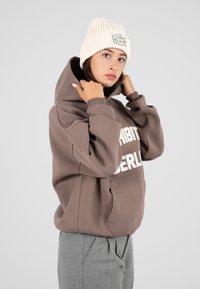 Giovane donna che sistema un berretto di lana color crema, indossando una felpa oversize marrone con testo bianco e pantaloni grigi su uno sfondo chiaro e neutro.