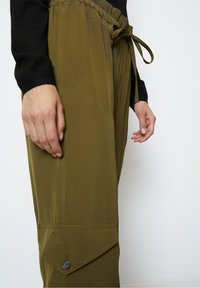 Olivově zelené kalhoty s elastickým pasem, stahovací šňůrkou a postranní kapsou s detailním patentkem. Hladká, lehká tkanina.