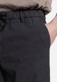 Mørkmønstret shorts med elastisk talje og snøre, der har en glat tekstur og sidelommer, delvist synlig hånd.