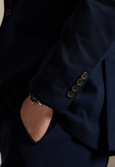 Man's hand in pocket of dark blue suit jacket with four buttons on cuff, wristwatch visible under sleeve.