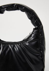 Sac à main rembourré noir avec une poignée arrondie et froncée. Fabriqué en matériau brillant, présente un détail de fermeture éclair argenté.