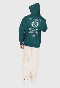 Person in einem dunkelgrünen Kapuzenpulli mit weißem Text und Emblem auf dem Rücken, beigen Cargo-Hosen und weißen Sneakers, mit dem Rücken zur Kamera und Kapuze übergezogen.