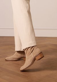 Stivaletti in suede beige con punta affusolata, tacco in legno basso e impuntura morbida e testurizzata. Indossati con pantaloni di colore chiaro.
