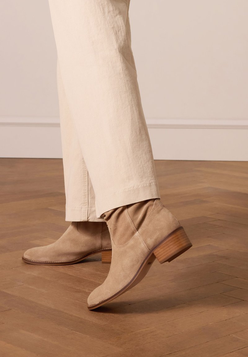Stivaletti in suede beige con punta affusolata, tacco in legno basso e impuntura morbida e testurizzata. Indossati con pantaloni di colore chiaro.