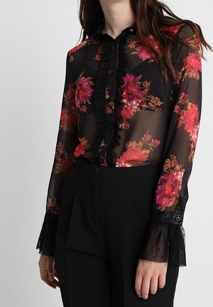 Vrouw die een doorschijnende zwarte blouse draagt met rood bloemmotief, gerimpeld voorpand en kanten manchetten, gecombineerd met zwarte hoge taillebroeken.