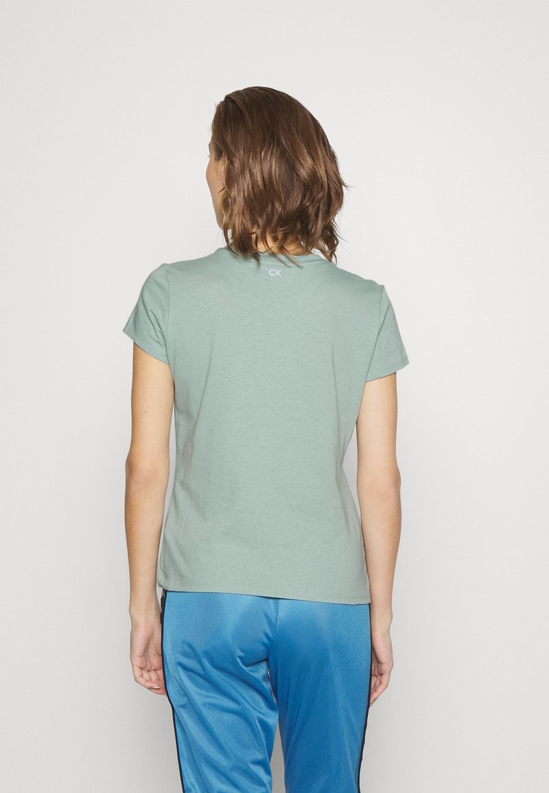 Camiseta de manga corta verde menta con cuello redondo y un sutil logo "CK" en la parte posterior. Tela suave con forma ajustada. Combinada con pantalones azules.