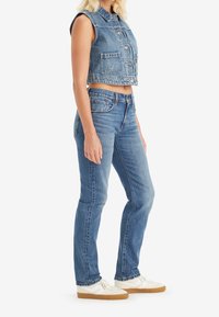 Chaleco recortado de mezclilla azul claro con diseño sin mangas, bolsillos frontales, combinado con jeans ajustados azul claro y zapatillas blancas con suelas marrones.
