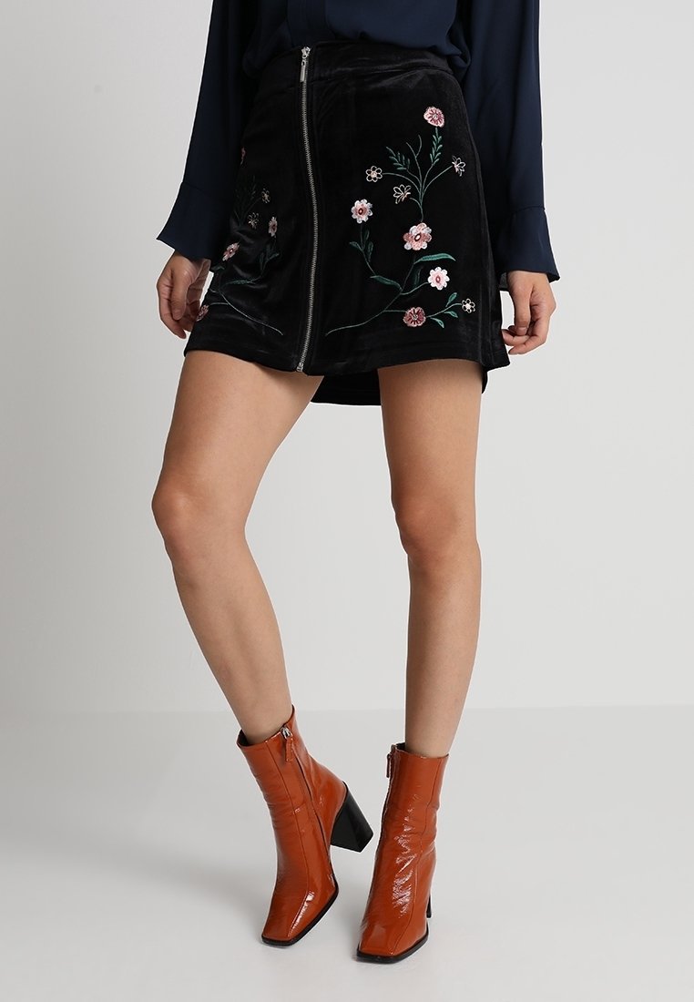 Zwarte fluwelen rok met bloemdecoratie, vooraan met rits en in a-lijnvorm. Gecombineerd met oranje hakschoenen tot enkel.