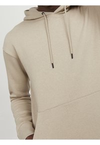 Sudadera beige con capucha hecha de una tela suave, con un bolsillo tipo canguro, cordones ajustables y puños de canalé. Textura suave.