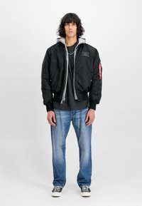Young man with curly hair wearing layered black bomber jacket, gray hoodie, black shirt, blue jeans, and black sneakers, standing on white background.